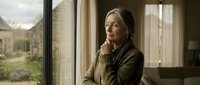 Mature woman with long gray hair and olive-toned skin, wearing a brown cardigan, gazing thoughtfully out a window with natural light streaming in, contemplative mood.