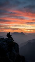 Hiker silhouette sitting on mountain cliff edge during dramatic sunset with colorful sky and flying birds over layered mountain ranges.