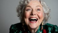 Joyful senior woman with silver curly hair wearing emerald green sequin dress and gold earrings expressing genuine happiness and laughter against light background.