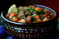 Hearty beef stew with tender meat chunks, carrots, potatoes in rich broth garnished with fresh cilantro and lime wedge served in traditional ceramic bowl.