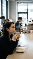 Young Asian woman enjoying coffee at modern cafe counter with blurred customers in background. Urban lifestyle and social dining concept.