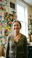 Smiling middle-aged Caucasian female teacher standing in elementary school classroom with colorful student artwork displayed on walls behind her.