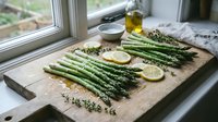 Fresh asparagus spears with lemon slices and herbs on wooden cutting board by kitchen window, ready for healthy cooking and meal preparation.