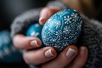 Decorative blue Easter egg with white floral folk art pattern held in feminine hands with french manicure against cozy gray knitted background.