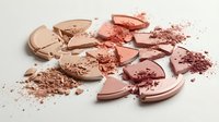 Broken coral pink makeup powder compacts scattered with crumbled cosmetic dust on neutral background for beauty and fashion projects.