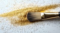 Professional makeup brush sweeping through scattered gold glitter powder on white surface, creating dynamic motion trail effect for cosmetic and beauty concepts.