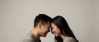 Young Asian couple sharing intimate moment with foreheads touching, smiling warmly in cozy sweaters against neutral background for relationship content.