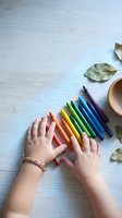 Child hands reaching for colorful crayons on white wooden table with autumn leaves and wooden bowl for creative art projects and educational activities.