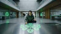 Young woman with dark hair and olive complexion, wearing olive-green jacket, interacting with glowing green holographic interface on floor in modern corridor. Technology, innovation, futuristic design.