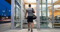 Athletic man entering modern fitness center through glass doors carrying gym bag ready for evening workout session.