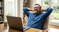 Relaxed mature businessman with beard stretching at desk in bright home office with laptop, expressing satisfaction and work-life balance success.