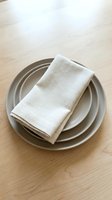 Overhead view of stacked neutral ceramic plates with folded cream linen napkin on light wood surface. Suitable for dining, hospitality, and table setting design projects.
