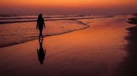 Woman in wetsuit walking along beach at sunset with reflection in wet sand, ideal for travel and lifestyle content.