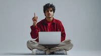 Young Hispanic man in red shirt sits cross-legged with laptop, raising finger in eureka moment against gray background. For business, education, and lifestyle projects.