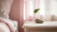 Cozy bedroom nightstand with a glass vase holding a green leaf and a stone inscribed hello in soft pink morning light.