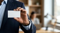 Businessman in dark suit holding blank white business card in office environment with colleagues working in background.