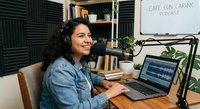Young Hispanic woman podcaster smiling while recording in home studio with laptop, microphone and acoustic foam panels for professional audio content creation.