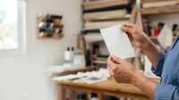Male artist in blue shirt holding blank white paper in creative studio workspace with shelves and art supplies.