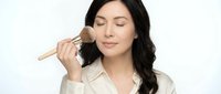 Young Hispanic woman applying makeup with brush, eyes closed in peaceful expression, wearing cream blazer on white background for beauty and cosmetics content.