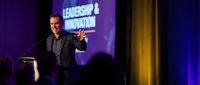 Middle-aged Caucasian businessman speaking at leadership conference with microphone and presentation screen in background with purple lighting.