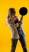 Young blonde woman holding black frying pan in casual outfit against bright yellow background for cooking and kitchen lifestyle concepts.