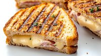 Grilled sandwich with melted cheese and ham on toasted bread, showing grill marks and herbs, cut diagonally to display melty interior on white surface.