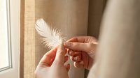Caucasian woman holding delicate white feather against neutral beige curtain background, symbolizing lightness, purity, and gentle spirituality for wellness and lifestyle content.