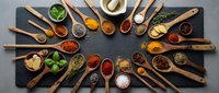 Colorful assortment of aromatic spices and herbs in wooden spoons arranged on dark slate stone background, top view flat lay.