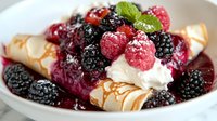 Fresh raspberries and blackberries top cream-filled meringue roll garnished with mint leaves and drizzled caramel sauce in white ceramic bowl, macro food photography.