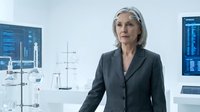Mature woman with gray hair and Caucasian ethnicity wearing dark business blazer in modern laboratory or research facility with scientific equipment and digital displays in background.