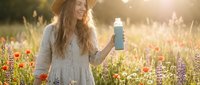 Young Caucasian woman with long wavy hair in straw hat holding reusable water bottle in wildflower meadow during golden hour sunset for eco-friendly lifestyle.