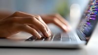 Close-up view of hands typing on laptop keyboard with programming code visible on blurred screen in background, representing software development and coding.