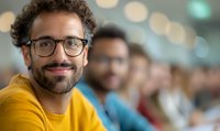 Young Hispanic man with glasses and beard wearing yellow sweater smiling confidently at camera. Blurred diverse group of people in background creates depth of field effect.
