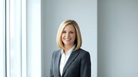 Professional blonde businesswoman in dark suit smiling confidently in modern office environment with natural lighting for corporate presentations.