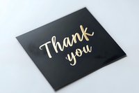 Elegant gold lettering "Thank you" message on black card against white background, modern calligraphy style text for greeting cards and appreciation notes.