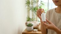 Young woman of East Asian descent holding white cosmetic bottle with blurred potted plants in background, suitable for skincare, beauty product marketing, wellness and natural cosmetics advertising.