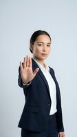Professional Asian businesswoman in navy suit making stop gesture with hand raised against white background for corporate presentations.