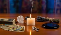 Mystical fortune telling setup with tarot cards, crystal skull, burning candle, sage bundle and scrying mirror on wooden table for spiritual guidance.