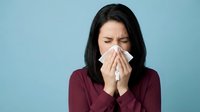 Young woman with dark hair and olive complexion, wearing burgundy top, blowing nose with tissue against blue background. Suitable for healthcare, wellness, and cold season campaigns.