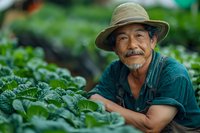 Senior Asian male farmer in straw hat and denim overalls smiling while working in organic vegetable garden with fresh green cabbage plants on sunny day.