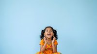 Young girl with dark curly hair and medium skin tone wearing orange shirt, expressing joy and excitement with open mouth and hands near face against blue background.