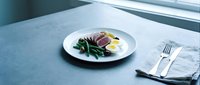Nicoise salad with seared tuna, green beans, olives, and boiled egg on a white plate, served on a gray stone surface with cutlery.