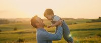 Father lifting happy toddler son in air during golden hour sunset in countryside field. Warm family bonding moment outdoors in rural landscape setting.