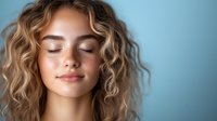 Young Caucasian woman with closed eyes and gentle smile, wavy blonde hair against blue background. Natural beauty portrait showing peaceful, serene expression.
