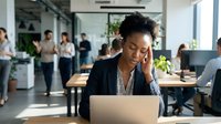 Stressed African American businesswoman experiencing headache while working on laptop in modern office with colleagues in background.