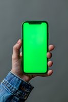 Hand holding modern smartphone with bright green screen display against gray background, wearing denim jacket sleeve. Mockup for app and mobile design.