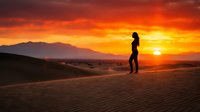 Woman standing on sand dunes at golden hour sunset, silhouetted against dramatic orange and yellow sky with mountains in background. Suitable for travel, wellness, and inspirational content.