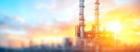 Industrial refinery chimneys against dramatic sunset sky with golden sunlight and blue atmosphere, panoramic view showing energy production facility.