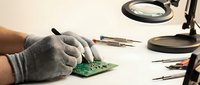 Technician in protective gloves repairing electronic circuit board with precision tools under magnifying lamp for professional electronics service.