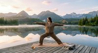 Young woman practicing yoga warrior pose on wooden dock at mountain lake during peaceful sunrise with perfect water reflections and forest landscape.